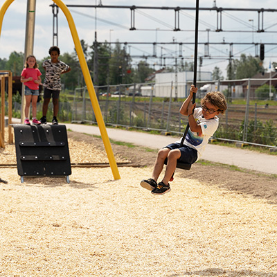 Ein kleines Kind benutzt eine Seilrutsche auf einem Spielplatz, es sitzt in der Mitte des Weges auf einem kleinen Sitz und hält sich am Seil fest.